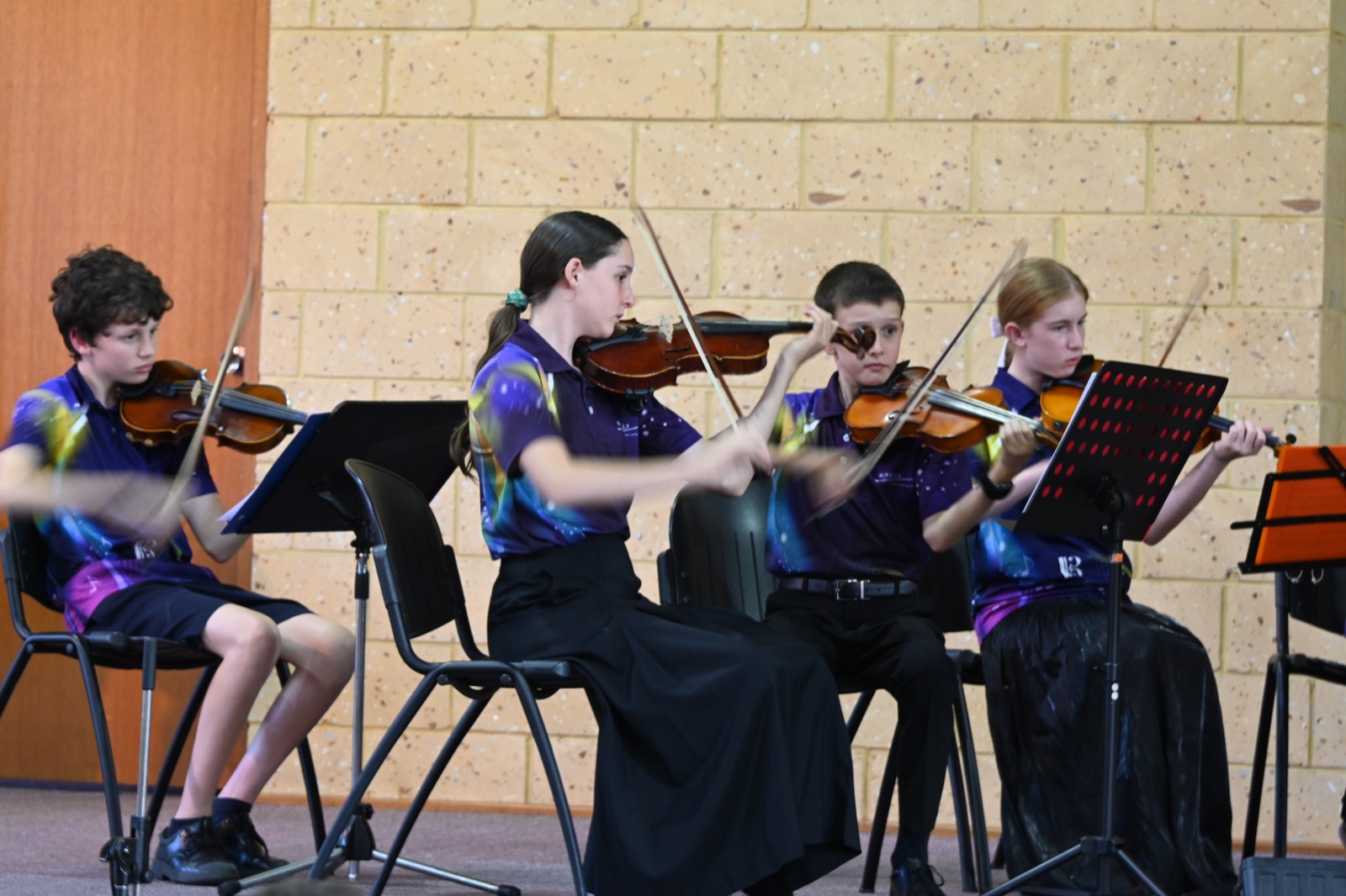 Four young violinists in MNCYO shirts performing in a row at a concert.